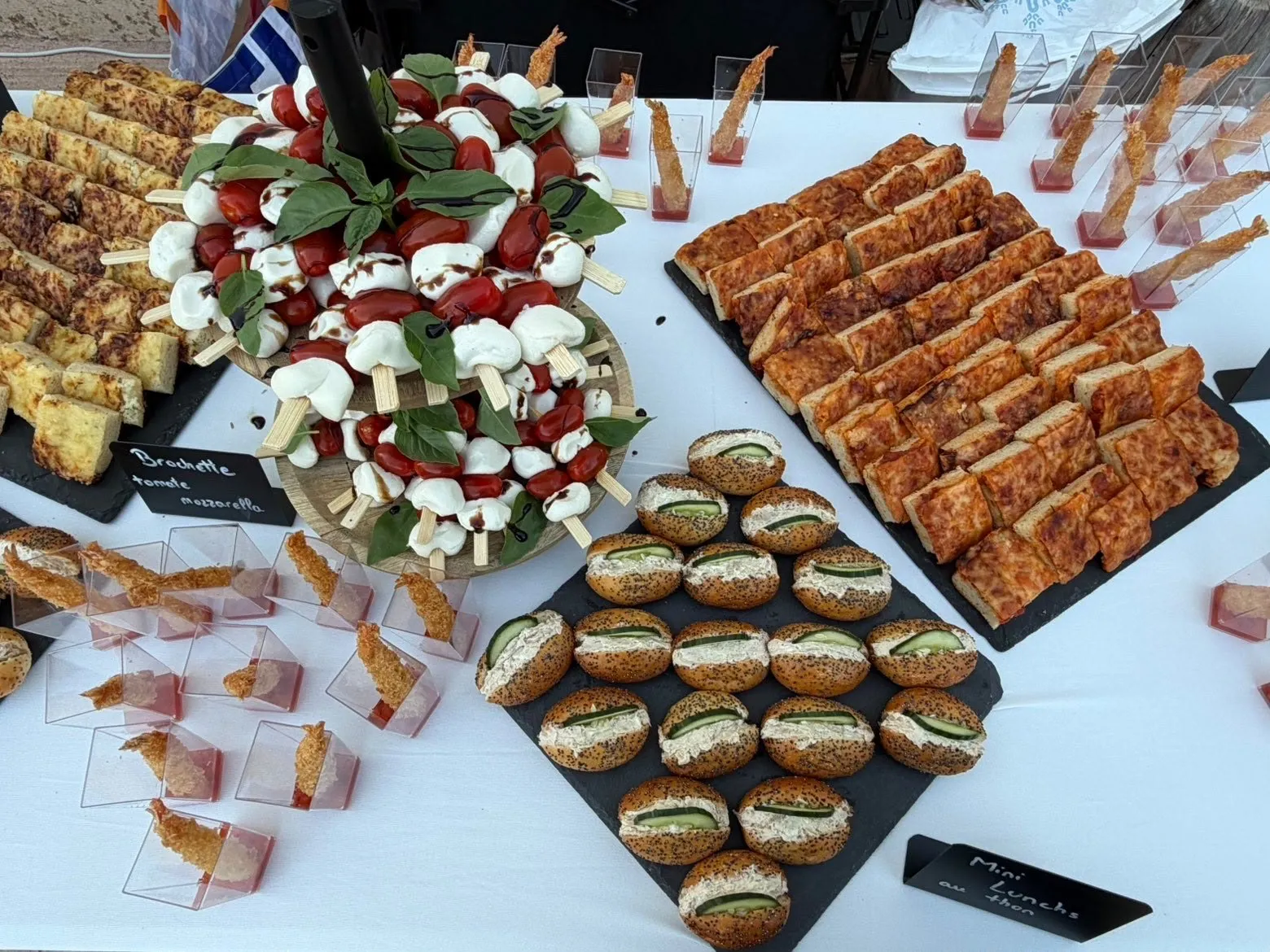 Buffet dressé en extérieur : brochettes tomate-mozzarella sur présentoir, pizza, mini lunchs au thon et verrines de crevettes croustillantes